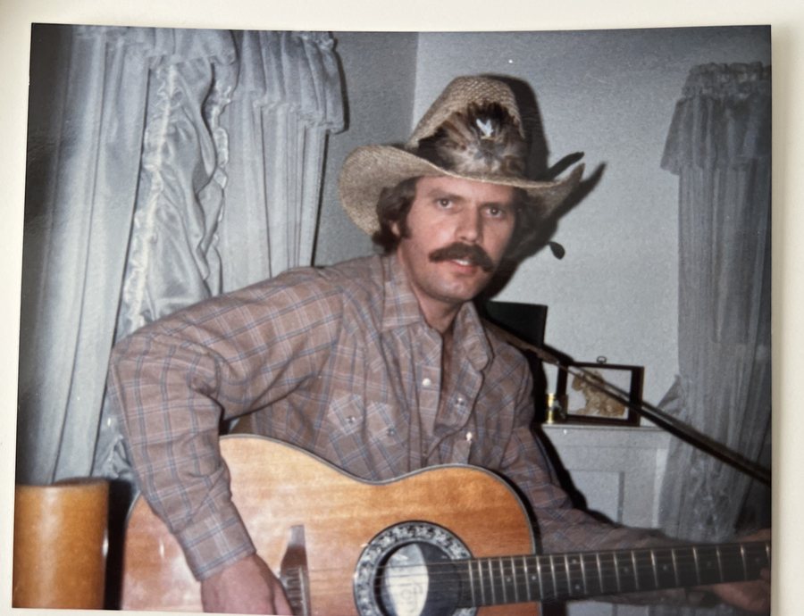 A young Geezer Jim in a plaid shirt and straw hat, guitar in hand