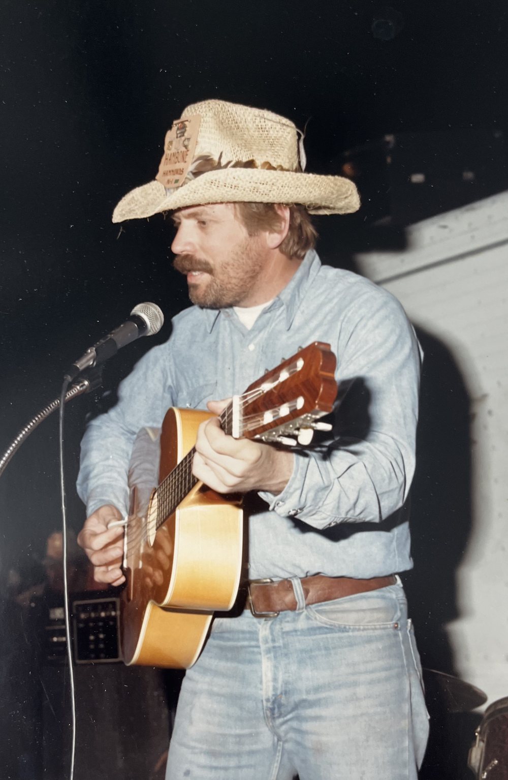 Geezer Jim on stage in a straw hat, singing at the mic with his guitar